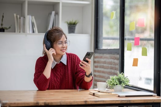 Asian female college student using laptop and phone with headphones while studying. Reading messages and greeting friends via video call.