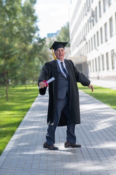 Old happy man in graduation gown jumping outdoors and holding diploma. Vertical