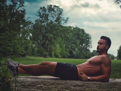 A muscular shirtless man laying on top of a rock in a field, looking up at the sky