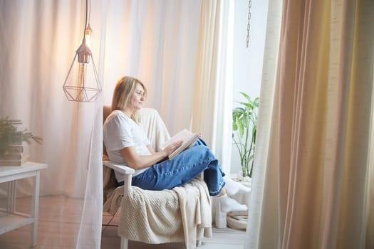 Young blonde woman with book on chair near window at home. Student girl is resting and reading textbook at home. Nice atmosphere. The concept of cozy home