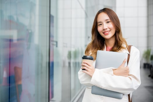 Beautiful portrait of a multitasking woman shows her walking against a beige wall with a takeaway coffee cup and a laptop in her hands. She smiling and confident, successfully work