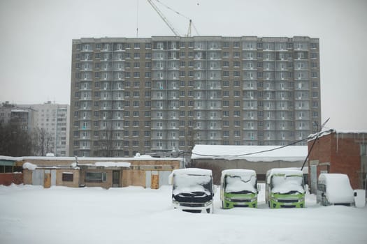 Cityscape in winter. View of construction site of house. Parking for buses. Snow-covered industrial zone.