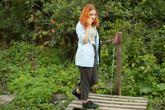 Girl pours water and talks on phone. Teenager in rural areas. Girl in village. Red hair.