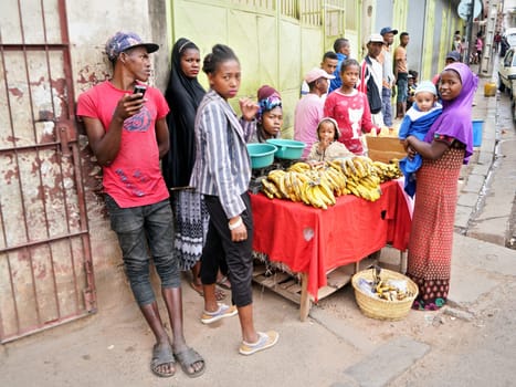 Antananarivo, Madagascar - April 24, 2019: Group of unknown Malagasy people standing near woman selling bananas on the street. Country is poor and locals usually cannot afford shopping in supermarkets