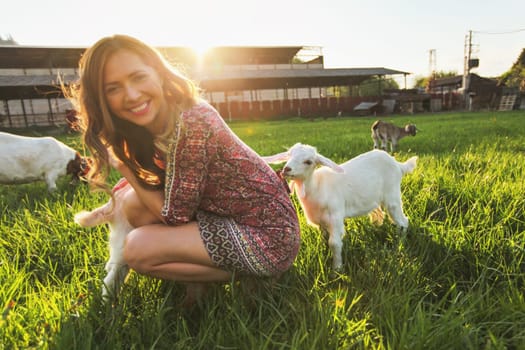 Young woman playing with goat kids on green spring meadow, smiling as animal chews her dress. Wide angle photo in strong sun backlight