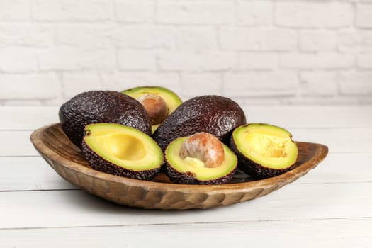 Ripe brown avocados ( hass - bilse variety ) some of them halved in wooden carved bowl, on white boards desk, brick wall background