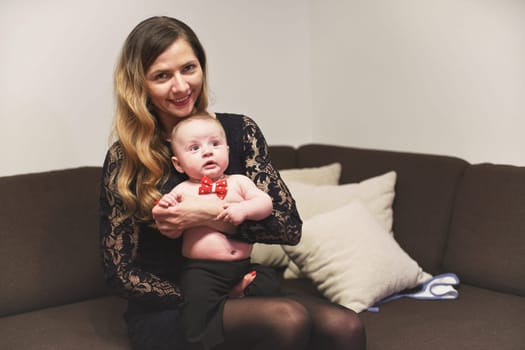 Young woman holding 4 months old infant baby boy with red bow tie on her hands, sitting on the couch