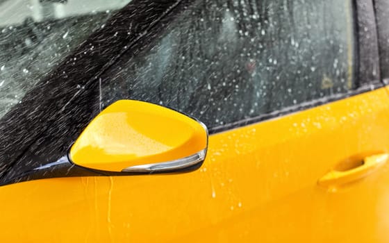 Front side mirror of bright yellow car washed in self serve carwash, water spraying over glass