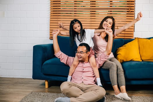 In their modern house an Asian family dad mom and daughter cuddle on a comfortable sofa sharing laughter and smiles during self-isolation. The essence of togetherness and happiness is evident.