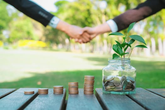 Sustainable money growth investment, glass jar filled with money saving and coin stack with businesspeople handshake in background as eco-friendly financial investment nurtured with nature. Gyre