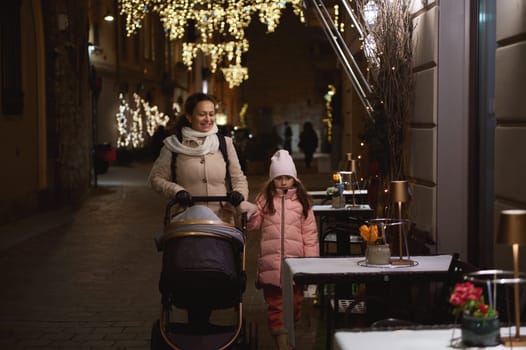 Authentic lifestyle portrait of a happy family of a mom holding the hand of her little daughter, pushing the baby pram, walking together on the street illuminated by festive lights in the night time