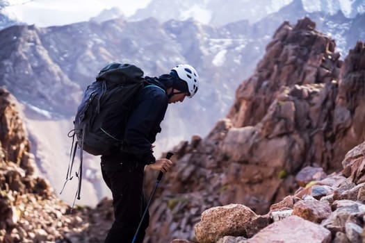 A young man traveler is engaged in mountaineering. In a helmet and with trekking poles, tourist climbs to the top, against the backdrop of a stunning view from the snow-capped mountains and rocks.