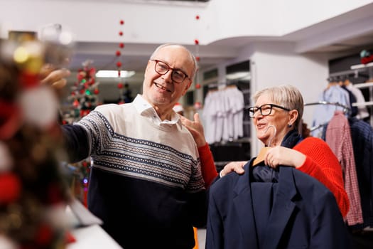 Senior couple searching clothing on videocall in retail store, trying to buy perfect fashion item measurement for christmas festivity. Two older people shopping for xmas gifts during holiday season.