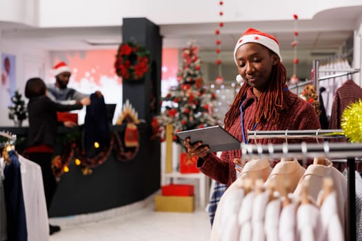 African american employee checking clothes stock, verifying list of trendy merchandise in shopping center, working on products inventory. Store manager counting clothing items on racks.