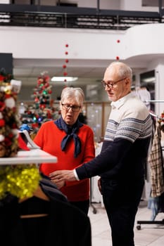 Senior persons browsing for jackets in shopping mall retail store, checking material of elegant clothing on sale. People looking to buy formal christmas gift for son, searching for quality blazer.