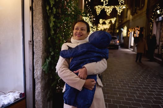 Happy elegant multi ethnic woman, loving mother carrying her baby, enjoying walk on the street in the night time, decorated with Christmas lights and glowing lanterns, smiling looking aside showcases