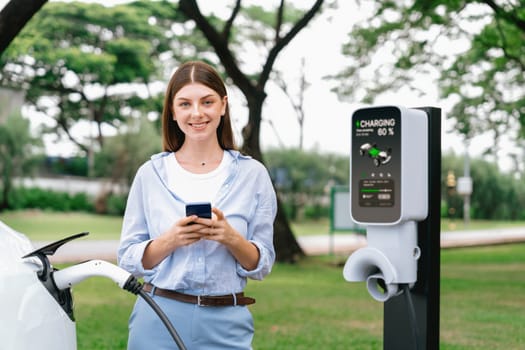 Young woman recharge EV electric vehicle battery from EV charging station and using smartphone online banking to pay for electricity in city park. Eco friendly vehicle travel with EV car. Exalt