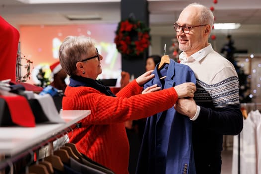 Senior couple examining blazers on racks in mall clothing store, looking for formal attire to wear on christmas dinner festivity. Elderly customers searching for elegant clothes in festive shop.
