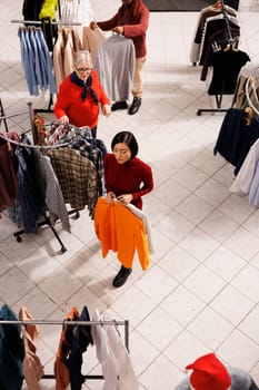 Diverse people shopping for clothes in retail store, looking for christmas presents and festive attire at mall. Women checking fabric of clothing items on hangers, buying trendy merchandise.