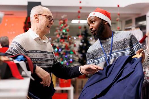 Male employee recommending clothes for senior customer, looking to buy formal outfit for christmas eve dinner. Elderly man asking worker for assistance to choose perfect fit clothing items.