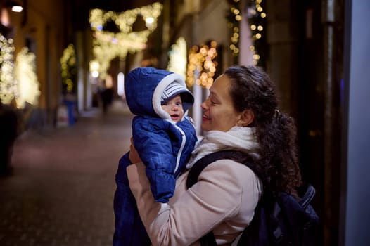 Portrait of a cheerful happy young woman mother smiles holding her baby boy, strolling the street of elegant Como city, enjoying the festive Christmas atmosphere, visiting fairground in the night