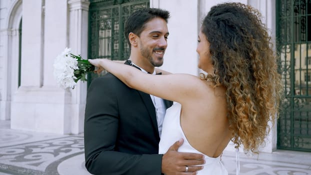 Newlyweds embrace with bouquet. Action. Beautiful young couple in stylish suits hugging in city. Stylish newlyweds embrace at church building.