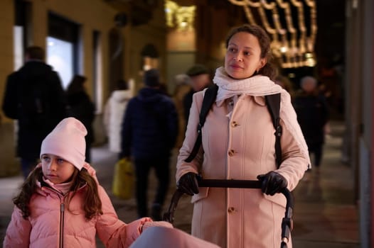 Mother with baby pram and her adorable little daughter strolling the street, enjoying festive atmosphere and Christmas funfair, visiting the traditional family market in December in the night time