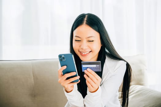 Happy woman holds smartphone and credit card shops online on sofa. Engaged in ordering banking and buying goods or services. Modern technology for convenient shopping.