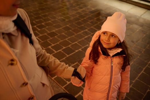 View from above of a Caucasian cute little child girl in warm winter clothes, holding the hand of her mother and cutely smiling looking up while strolling together the street in the night time