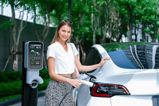 Young woman travel with EV electric car charging in green sustainable city outdoor garden in summer. Urban sustainability lifestyle by green clean rechargeable energy of electric BEV vehicle innards