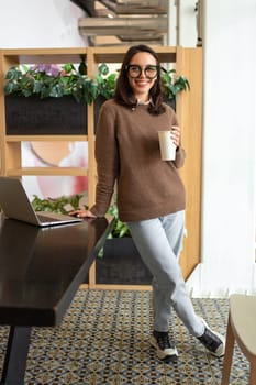 Woman in glasses poses smiling standing at workplace with cup coffee. Woman dressed in smart-casual is standing leaning on table and smiling. Female stands by work laptop happy to be relaxing