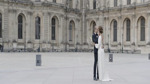 Newlyweds with an antique building on the background. Action. Beautiful bride and groom look each other and kiss