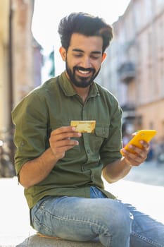 Indian man using credit bank card smartphone while transferring money, purchases online shopping, order food delivery, booking hotel room, town lifestyles. Arabian guy sitting in city street. Vertical