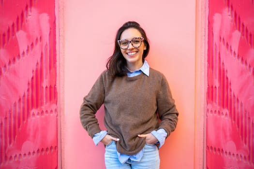 Woman smiling and poses for photo near pink wall in street. Female standing dressed in casual style smiling and holding hands in pants. Portrait woman posing stands in street with pink background