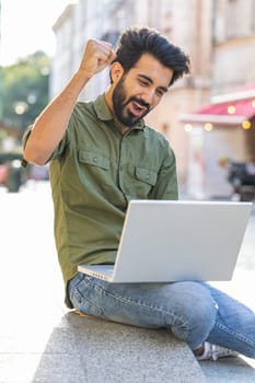 Happy indian man sitting in city street looking at laptop celebrating success, win message, lottery jackpot victory, giveaway online, playing game. Excited arabian guy get online good news. Vertical