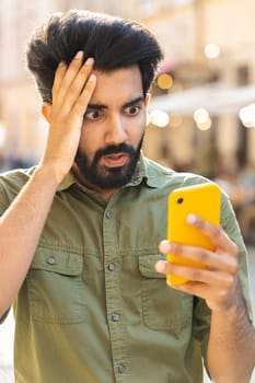 Indian bearded man use smartphone typing browsing, loses becoming surprised sudden lottery results, bad news, fortune loss, fail. Guy walking in urban city street outdoors. Town lifestyles. Vertical