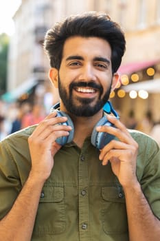 Joyful relaxed overjoyed indian man choosing, listening favorite energetic disco rock n roll music dancing outdoors. Guy taking off wireless headphones looking at camera in urban city street. Vertical