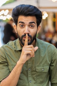 Shh be quiet please. Indian man presses index finger to lips makes silence hush gesture sign do not tells gossip secretoutdoors. Arabian guy walking in urban city street. Town lifestyles. Vertical