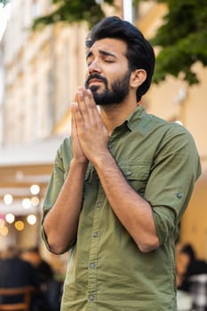 Bearded religion indian man praying with closed eyes to God asking for blessing, help, forgiveness outdoor. Adult guy clasping hands wishing luck in urban sunny city street. Town lifestyle. Vertical