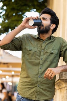 Happy bearded indian young man enjoying morning coffee hot drink, town lifestyles outdoors. Relaxing, taking a break. Smiling arabian muslim adult guy standing in urban city center street. Vertical