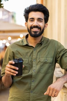 Happy bearded indian young man enjoying morning coffee hot drink, town lifestyles outdoors. Relaxing, taking a break. Smiling arabian muslim adult guy standing in urban city center street. Vertical