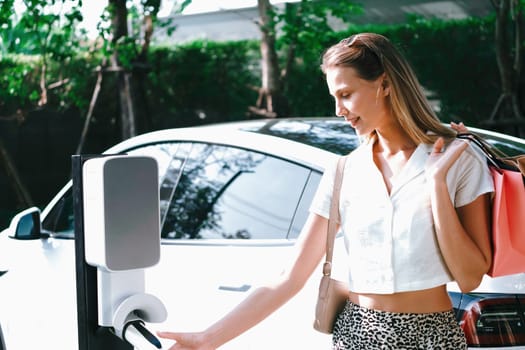 Young woman travel with EV electric car charging in green sustainable city outdoor garden in summer. Urban sustainability lifestyle by green clean rechargeable energy of electric BEV vehicle innards