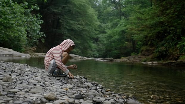 Boy child squatting in front of a river and collecting stones near the water. Creative. Kid exploring nature on a summer day