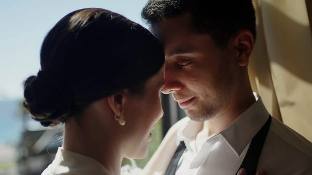 Cute newlywed couple embracing by the window with the beach behind. Action. Bride and groom in love by the sea