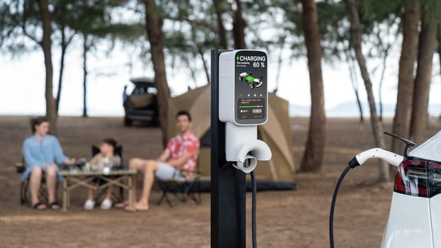 Alternative family camping trip traveling by the beach with electric car recharging battery from EV charging station with blurred family enjoying the seascape campsite background. Perpetual