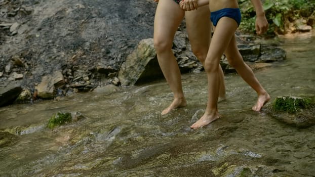 Legs of boy and mother in swimsuit walking along rocks in cold stream. Creative. Hiking in wild nature