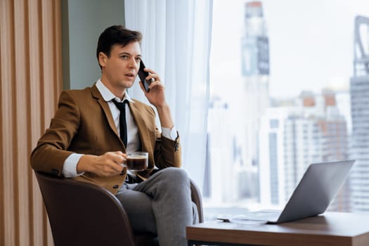 Closeup of handsome businessman making phone call with manager while sitting near window with skyscraper view. Executive manager talking working by using phone and laptop. Look aside. Ornamented.