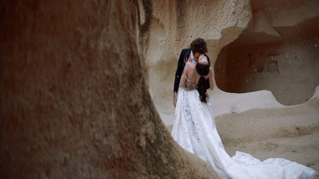Stylish beautiful bride and groom under falling snow near rocks or caves. Action. Wedding video shooting in cappadocia