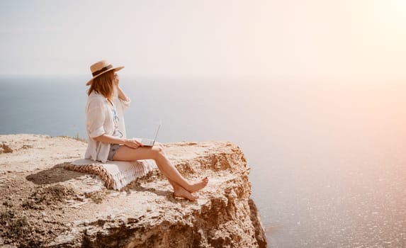 Successful business woman in yellow hat working on laptop by the sea. Pretty lady typing on computer at summer day outdoors. Freelance, travel and holidays concept.