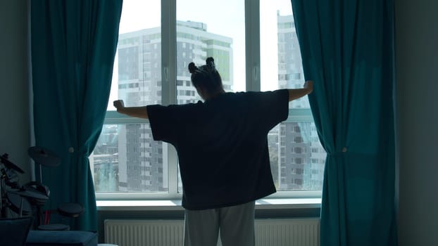 Rear view of a woman stretching while standing by the window. Media. Young girl in a bedroom in the morning